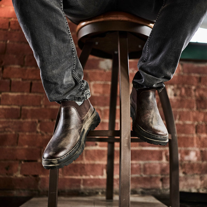 Person wearing brown leather boots and jeans sitting on a wooden stool against a brick wall.